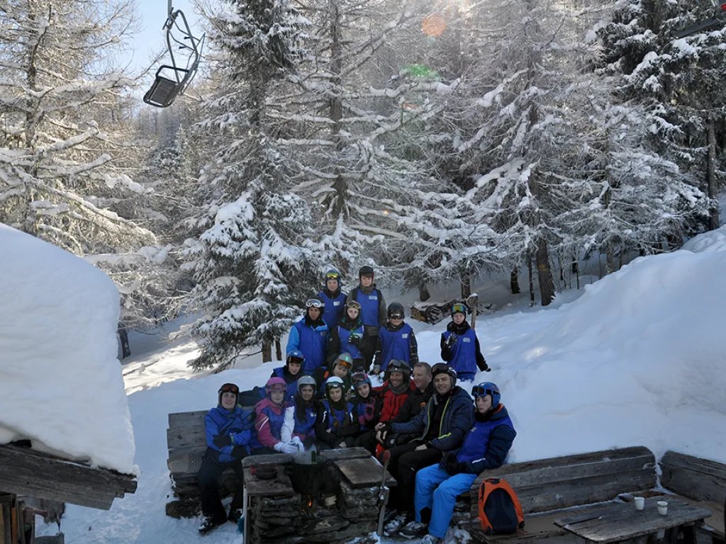 Gruppe sitzt an Holztischen vor einer Hütte im verschneiten Wald.